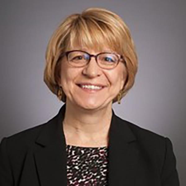 Professional image of a woman wearing a floral shirt, black blazer, and black reading glasses, sitting against a grey background.